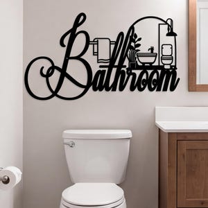 May include: A black metal wall sign featuring the word Bathroom in a cursive script. The design incorporates a line art illustration of a towel on a rack, a potted plant, a bathtub, and a toilet, all integrated into the lettering structure.