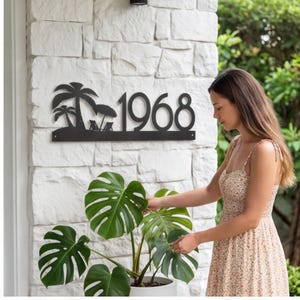 May include: Black metal address sign with the number 1968, palm trees, and beach chairs. The sign is mounted on a white brick wall. A woman in a floral dress is tending to a large green plant.