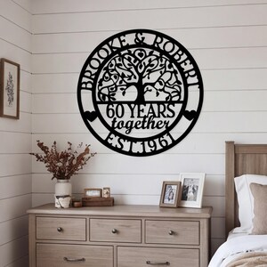Puede incluir: Un letrero de pared circular de metal negro con un diseño de árbol cortado con láser. El texto dice BROOKE & ROBERT, 60 YEARS together y EST. 1961. Dos pequeñas formas de corazón están situadas a los lados de la barra horizontal central.