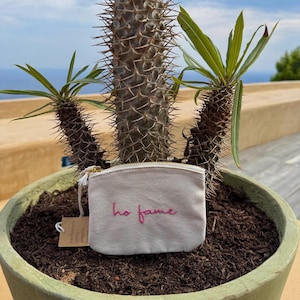 May include: A small, off-white canvas pouch with a gold zipper and a tassel. The phrase "ho fame" is embroidered in pink. The pouch is set in a green pot with a cactus and other plants, with a blue sky in the background.
