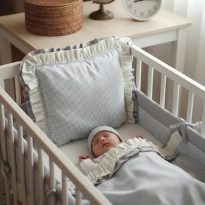 Può includere: Un bambino che dorme in una culla bianca con un cuscino e una coperta a quadretti grigi e bianchi. Il cuscino e la coperta hanno bordi arruffati bianchi. Il bambino indossa un cappello grigio e un body bianco.