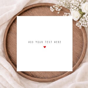 May include: A white square card with the words "ADD YOUR TEXT HERE" and a small red heart. The card sits on a wooden tray, with white flowers and fabric in the background.