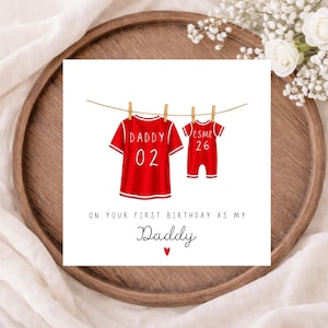 May include: A birthday card featuring a red jersey and baby onesie suspended from a clothesline. The jersey displays "DADDY 02" and the onesie reads "ESME 26". The card's message is "ON YOUR FIRST BIRTHDAY AS MY Daddy" with a heart.