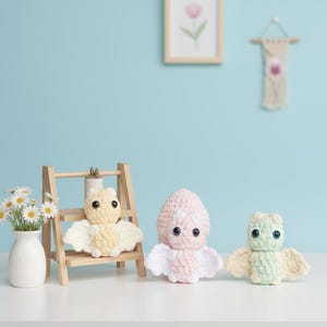 May include: A collection of handmade crochet toys, including a yellow bat on a wooden ladder, a pink and white octopus, and a green and yellow owl. A vase of daisies sits nearby. The toys are on a white surface against a light blue wall.