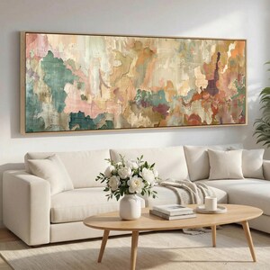 May include: Large abstract painting in a rectangular wooden frame. The artwork features a textured surface with a palette of muted colors, including greens, pinks, and browns. The painting hangs above a white sofa and a wooden coffee table.