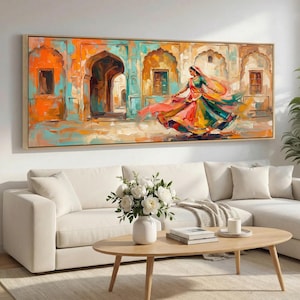 Puede incluir: Una pintura al óleo sobre lienzo representa a una mujer bailando con un vestido colorido frente a un telón de fondo arquitectónico indio. La obra de arte presenta tonos vibrantes de naranja, turquesa y amarillo, enmarcada en madera natural. El cuadro está sobre un sofá blanco.
