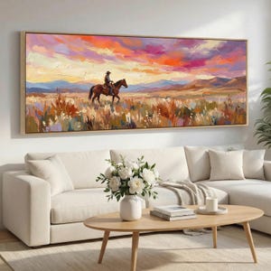 May include: A rectangular framed painting depicts a cowboy on horseback in a field at sunset. The sky is a vibrant mix of orange, pink, and purple hues. The artwork is set in a landscape with mountains in the background, and the frame is a light wood color.