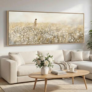 May include: A large rectangular framed canvas art piece depicts a figure walking through a field of white flowers. The artwork has a soft, muted color palette with a beige wooden frame. The scene is serene and peaceful.