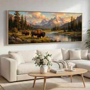 May include: A framed landscape painting showing two bison by a river in a mountain valley. The artwork uses a warm colour palette with a sunset sky, snow-capped mountains, and lush greenery. The frame is a light wood colour.