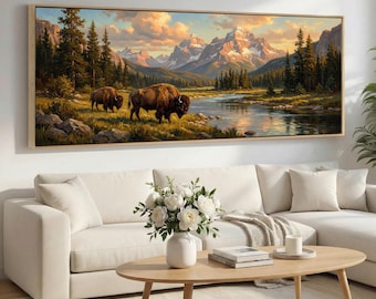 May include: A framed landscape painting depicts two bison near a river in a mountain valley. The artwork features a warm color palette with a sunset sky, snow-capped mountains, and lush greenery. The frame is a light wood color.