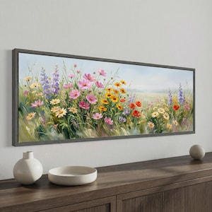 May include: A framed landscape painting depicts a vibrant field of wildflowers. Pink cosmos, yellow daisies, red poppies, and purple wildflowers bloom in a meadow. The artwork is encased in a gray wooden frame, creating a serene and colorful scene.