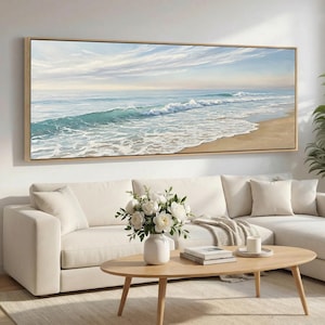 May include: A large rectangular framed painting depicts a serene ocean scene. The artwork showcases a sandy beach, turquoise waves, and a light blue sky with wispy clouds. The painting is displayed above a white sectional sofa and a wooden coffee table.