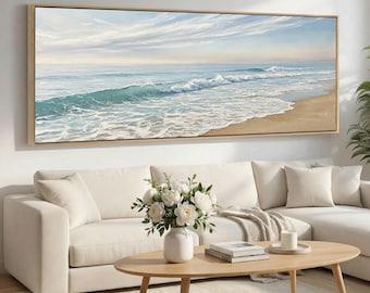 May include: A large rectangular framed painting depicts a serene ocean scene. The artwork showcases a sandy beach, turquoise waves, and a light blue sky with wispy clouds. The painting is displayed above a white sectional sofa and a wooden coffee table.