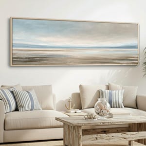 May include: A large rectangular framed canvas art piece depicting a serene coastal landscape. The artwork features a muted color palette of blues, tans, and grays, evoking a sense of calm. The painting is displayed above a beige sofa.