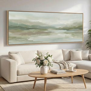 Könnte beinhalten: Abstrakte Landschaftsmalerei in einem rechteckigen Holzrahmen. Das Kunstwerk zeigt weiche, gemischte Farben in Grün, Beige und Weiß, die ein Gefühl der Ruhe hervorrufen. Das Gemälde hängt über einem weißen Ecksofa, mit einem Holztisch und einer Vase mit weißen Blumen.