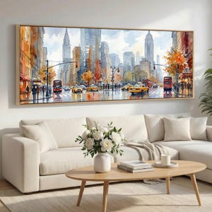 May include: A watercolor painting of a New York City street scene, featuring yellow taxis, red double-decker buses, and tall buildings. The artwork is framed in a light wood tone and hangs above a white sofa. The painting captures the city's architecture and autumn colors.