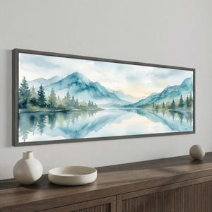 Könnte beinhalten: Ein gerahmtes Aquarellgemälde zeigt eine ruhige Berglandschaft. Das Kunstwerk zeigt blaue und grüne Berge, die sich in einem ruhigen See spiegeln, mit Bäumen am Ufer. Das Gemälde ist in einem dunkelgrauen rechteckigen Rahmen eingefasst.