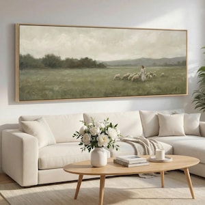 May include: A framed landscape painting shows a shepherd with sheep in a green field under a cloudy sky. The artwork is in a light-coloured wooden frame. The painting hangs above a white sectional sofa and a wooden coffee table.