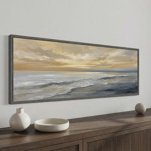 Può includere: Un'opera d'arte su tela incorniciata, di forma rettangolare e allungata, raffigurante un paesaggio marino. L'opera presenta una palette di colori tenui di blu, grigi e oro, con un cielo nuvoloso e dolci onde. La cornice è grigio scuro. Vaso e ciotola decorativi in ceramica bianca su una superficie di legno.