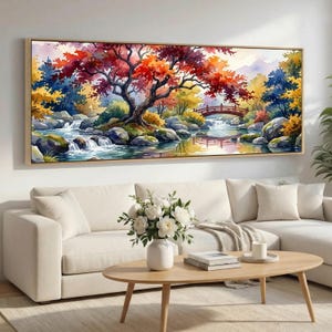 Pode incluir: Uma grande pintura retangular em aquarela de uma paisagem serena. Uma vibrante árvore vermelha e laranja domina o centro, com uma pequena ponte e água corrente. A obra de arte é emoldurada em madeira clara e pendurada acima de um sofá branco.