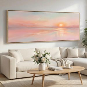 May include: A long, rectangular canvas artwork showing a sunset over water. The painting uses soft pinks, oranges, and blues, and is framed in gold. It hangs above a white sofa and a wooden coffee table, creating a calming atmosphere.
