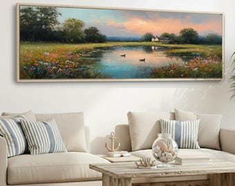 May include: A framed landscape painting depicts a serene pond scene with ducks, wildflowers, and a distant house. The artwork features a vibrant sky with sunset hues, lush greenery, and a light-colored wooden frame. The painting is displayed above a beige sofa.