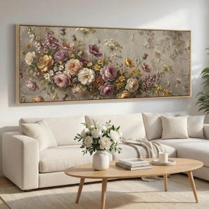 Op de afbeelding: Een rechthoekig bloemenschilderij in een licht houten frame. Het kunstwerk toont een verscheidenheid aan bloemen in tinten roze, geel en paars tegen een getextureerde, neutrale achtergrond. Het schilderij wordt boven een witte bank in een woonkamer getoond.