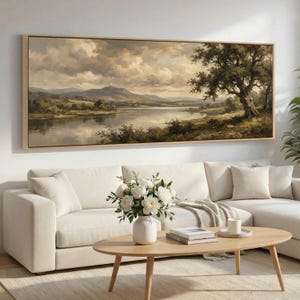 May include: A framed landscape painting depicts a serene lake scene with mountains in the background and a large tree on the right. The artwork is in a neutral color palette, with a light wood frame. A white sofa and coffee table are in the foreground.