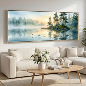 May include: A framed landscape painting shows a tranquil lake scene with trees and a sunset. The artwork has a light wood frame and is displayed above a white sofa. The painting's colours include blues, greens, and yellows, creating a peaceful atmosphere.