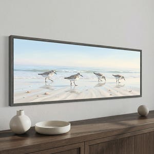 May include: A framed rectangular artwork depicts four sandpipers on a sandy beach with gentle waves under a light blue sky. The artwork is encased in a gray wooden frame, creating a serene coastal scene.