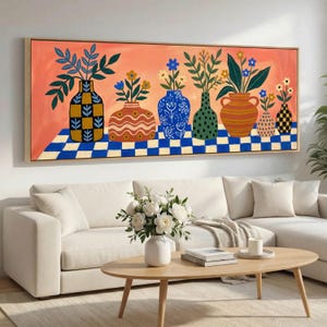 Op de afbeelding: Een ingelijst schilderij toont een rij kleurrijke vazen, elk met bloemen en bladeren. De vazen staan tegen een perzikachtergrond en een blauw-wit geblokt oppervlak. Het schilderij hangt boven een witte bank en een houten salontafel.
