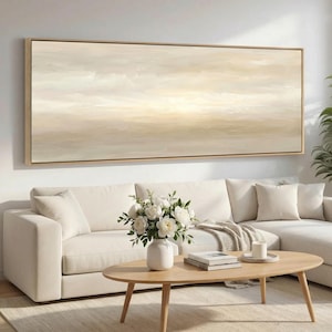 May include: A large, rectangular abstract painting in a light beige and cream colour palette, framed in light wood. The painting hangs above a white sectional sofa, with a wooden coffee table and a vase of white flowers in front.