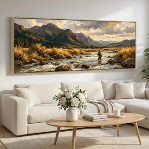 Può includere: Un dipinto di paesaggio incorniciato raffigura una serena scena fluviale con una persona che pesca. L'opera d'arte presenta una catena montuosa, erbe dorate e un cielo nuvoloso. Il dipinto è esposto sopra un divano bianco e un tavolino di legno.