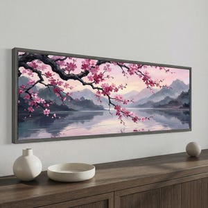 Pode incluir: Uma pintura de paisagem emoldurada retrata uma cena serena de um lago e montanhas sob um céu suave, rosa e roxo. Ramos de cerejeira com flores rosas se estendem na cena. A moldura é cinza suave.