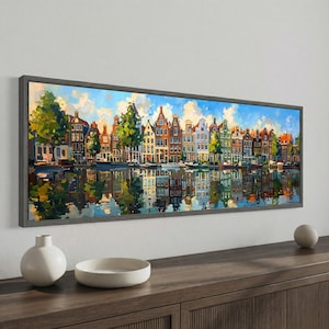 May include: A framed painting depicts a canal scene with colorful buildings and their reflections in the water. The artwork features a blue sky with clouds, trees, and boats, creating a vibrant and detailed landscape.