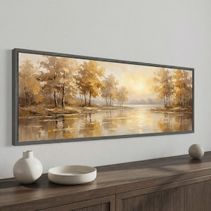 May include: A framed landscape painting shows a river scene with trees in gold and brown hues. The artwork reflects the sunlight, creating a warm atmosphere. The painting is displayed above a wooden cabinet. The frame is dark grey.