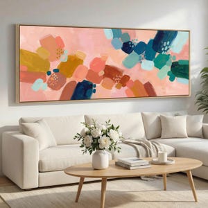 Op de afbeelding: Abstracte kunstafdruk in een rechthoekige houten lijst. Het kunstwerk toont penseelstreken in tinten roze, blauw, groen, geel en bruin. Het schilderij hangt boven een witte bank en een houten salontafel.