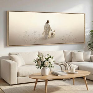 Op de afbeelding: Een ingelijste canvas print toont een figuur in witte gewaden met een lam, tegen een zachte, neutrale achtergrond. Het kunstwerk wordt tentoongesteld boven een witte bank, met een houten salontafel en een vaas met witte bloemen.