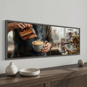 May include: A panoramic framed wall art print depicting a barista pouring steamed milk from a copper pitcher into a ceramic mug to create latte art. The scene features a professional espresso machine in the background and a dark apron.