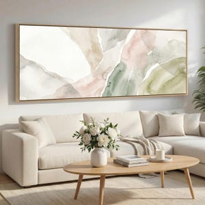 Może przedstawiać: Abstrakcyjny obraz akwarelowy w prostokątnej drewnianej ramie. Dzieło przedstawia miękkie, mieszane kolory bieli, różu, zieleni i beżu. Obraz jest wystawiony nad białą sofą z stolikiem kawowym i wazonem białych kwiatów.