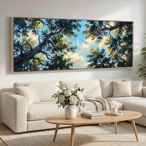 May include: An oil painting depicting a view upwards through a forest canopy, with a blue sky and clouds. The artwork is framed in a light wood frame and hangs above a white sofa. The painting's colours include greens, blues, and yellows.