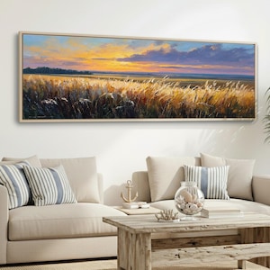 Pode incluir: Pintura de paisagem de um campo de capim alto ao pôr do sol. A obra apresenta tons dourados quentes na grama e no céu, com uma moldura de madeira. A pintura é exibida acima de um sofá bege com almofadas listradas.