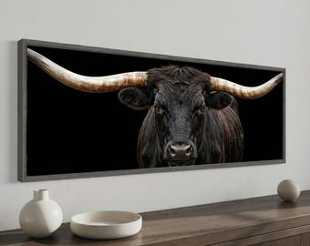 May include: A framed canvas print featuring a close-up of a black bull with large, curved horns against a black background. The bull's fur is dark brown, and the horns are a light tan color. The frame is a dark gray.