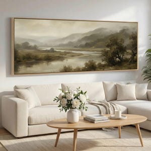 May include: A framed landscape painting depicts a serene river scene with rolling hills and trees in muted tones of brown and green. The artwork hangs above a white sofa, complemented by a wooden coffee table and a vase of white flowers.