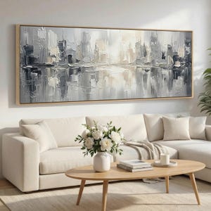 Op de afbeelding: Een groot, horizontaal rechthoekig wandkunstwerk met een abstract stadsgezicht in grijstinten, wit en beige. Het kunstwerk toont dikke, getextureerde penseelstreken en paletmesstreken, ingelijst in een lichtgekleurde houten lijst.