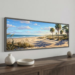 May include: A panoramic framed wall art print depicting a coastal beach scene with golden sand, tall dune grasses, and several palm trees under a bright blue sky with scattered white clouds. The ocean waves meet the shore in the distance.