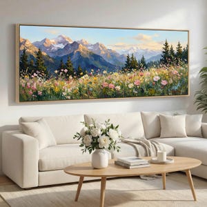 May include: A long landscape painting depicting a mountain range with snow-covered peaks, green forests, and a colourful meadow of wildflowers in pink, yellow, and white. The artwork is framed in light wood and hangs above a white sofa.