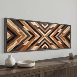 Può includere: Opera d'arte geometrica da parete in legno con cornice grigia. L'opera presenta un motivo simmetrico di assi di legno angolate nei toni del marrone, nero e beige. Il pezzo è rettangolare e montato su una parete bianca, sopra un mobile in legno.