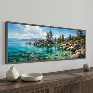 Op de afbeelding: Een ingelijst landschapsschilderij toont een serene meerlandschap. Het kunstwerk toont helder turquoise water, rotsachtige oevers en groenblijvende bomen onder een helderblauwe lucht met wollige wolken. Het frame is een gedempt grijs.