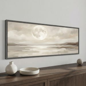 Op de afbeelding: Landschapsschilderij met volle maan boven water, in neutrale tinten, omlijst door donkergrijs hout. Een witte keramische vaas en kom staan op een houten oppervlak. Het kunstwerk roept een gevoel van sereniteit op.